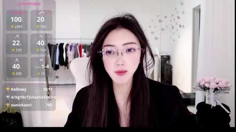 Video of an East Asian woman with long black hair and glasses, wearing a black top, sitting in a modern room with a mirror, plants, and clothes on a rack in the background.