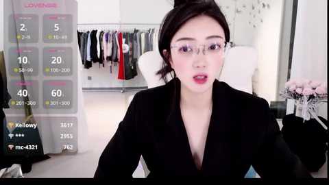 Video of an East Asian woman with fair skin, dark hair, and large glasses, wearing a black blazer, sitting in a modern, minimalist room with a calendar overlay.