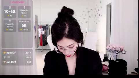 Video of a woman with dark hair in a bun, wearing glasses and a black top, standing in a modern, minimalist room with a mirror, flowers, and clothes in the background.