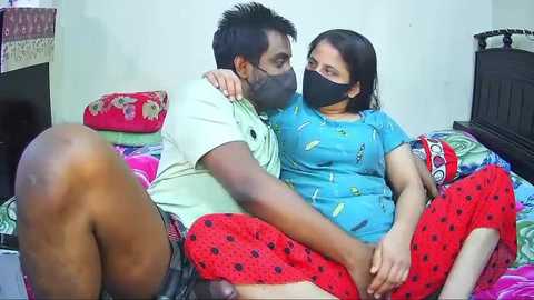 Video of a man and woman, both wearing face masks, sitting closely on a colorful bed, man in a yellow shirt and shorts, woman in blue shirt and red pants.