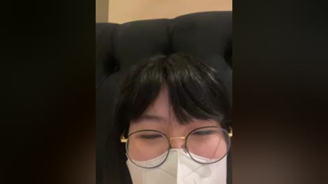 Video of a person with short black hair and round glasses, wearing a white mask, leaning back on a black, tufted leather sofa, with a blurred, neutral-colored background.