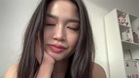 Video of a young Asian woman with long, straight black hair, light skin, and closed eyes, resting her chin on her hand, smiling softly. Background shows a white bookshelf with stuffed toys and decor.
