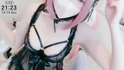 A young, pale-skinned woman with pink hair wears a black lace lingerie dress, white face mask, and black choker. She poses indoors, camera angled low, with a timestamp \"21:23 12/14 Sun\" in the corner.