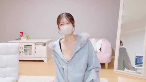 Video of an East Asian woman in a light blue, fluffy bathrobe, wearing a white face mask, standing in a minimalist, modern living room with white walls, a white couch, and a pink backpack.