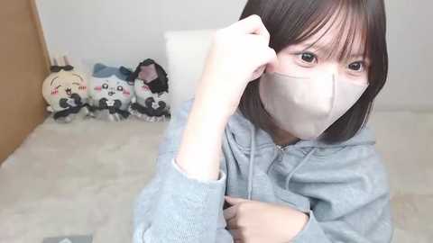 A video of an East Asian woman with short brown hair, wearing a gray hoodie and face mask, adjusting her hair. Background features plush toys and a white wall.