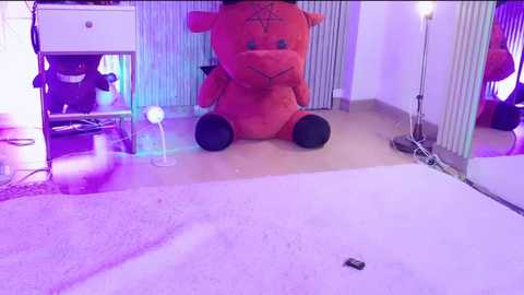 A video of a cozy room with a large, plush, red teddy bear on a cream carpet, illuminated by purple and blue lights. The background shows a white dresser, a chair, and a lamp.