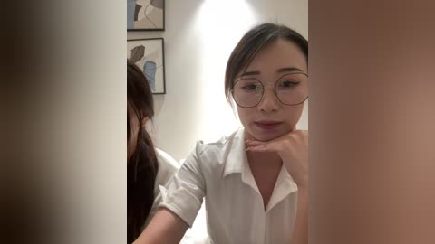 Video of an Asian woman with fair skin, glasses, and long dark hair, wearing a white shirt, leaning against a beige wall. She is in a modern, minimalist room with abstract art on the wall.