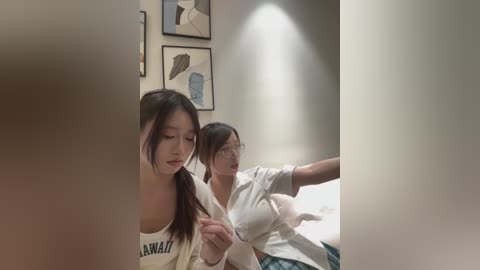 Video of two Asian women in a dimly-lit hallway, one wearing a white shirt and glasses, the other in a white blouse, both looking down. Background features abstract art and a glowing ceiling light.