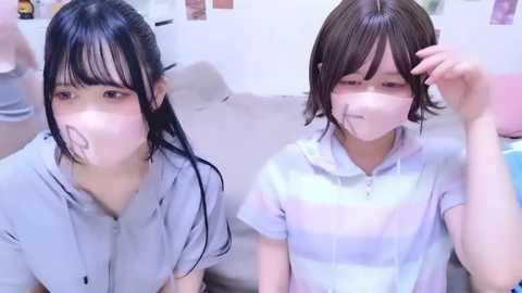 Media: Video of two young East Asian women in matching white face masks, wearing striped shirts, seated on a couch, with a soft, blurred background.