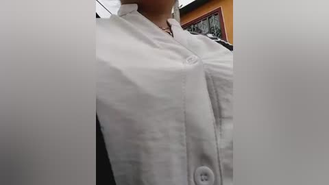 Video of a woman wearing a white chef's jacket with a large, prominent button, visible from the neck down. Background shows an orange wall and a framed picture.
