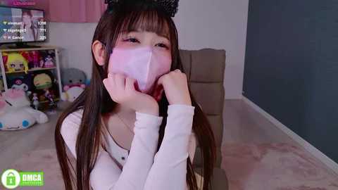 Video of a young Asian woman in a white long-sleeve shirt, wearing a pink face mask, sitting on a beige chair. Background features a pink wall, stuffed toys, and a desk.