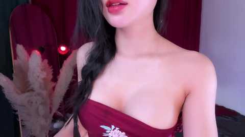 Video of an Asian woman with fair skin, long black hair, and full lips, wearing a red strapless top with floral embroidery, standing against a red curtain backdrop.