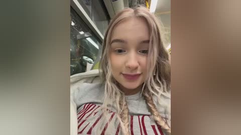 Video of a young woman with long, pastel pink hair and braided pigtails, wearing a striped sweater, standing inside a grocery store.