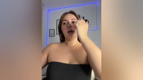 Video of a fair-skinned, busty woman with light brown hair, wearing a strapless black top, applying makeup in a modern, dimly-lit bedroom with a blue LED strip light.