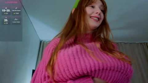 Media: Video of a smiling woman with long red hair, wearing a bright pink, ribbed sweater, standing in a dimly lit room with a digital clock displaying 15:30.