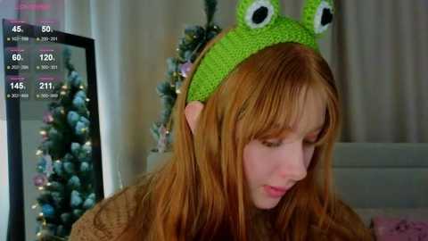 Video of a young woman with long, red hair, wearing a green frog hat and a brown sweater, looking down in a cozy room with a Christmas tree and digital stats overlay.