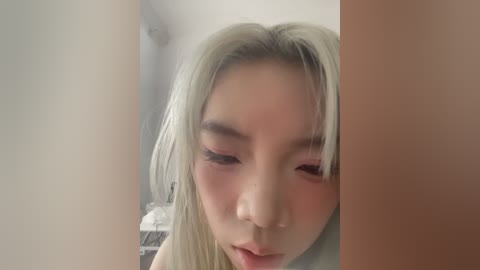 Video of an Asian woman with long, platinum blonde hair, fair skin, and full, pink lips, partially obscured by a beige pillar. Background shows a dimly lit room with a white wall and indistinct furniture.