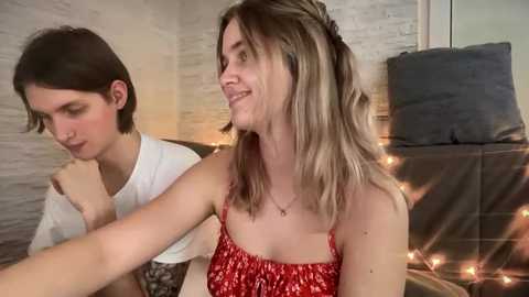 Video of two women in a cozy bedroom setting: one with short dark hair, wearing a white shirt, and the other with long blonde hair, in a red floral dress, smiling.