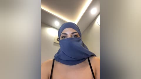 Video of a woman with fair skin and dark hair, wearing a blue face mask, black spaghetti straps, and a matching blue headscarf, indoors with beige walls and recessed ceiling lights.