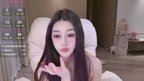 Video of a young East Asian woman with long black hair, fair skin, and a slender physique, wearing pink lipstick. She is lying on a white bed, smiling and blowing a kiss.