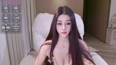Video of an Asian woman with long, straight black hair, light skin, and full lips, wearing a pink lace lingerie top, sitting on a white chair in a modern bedroom.