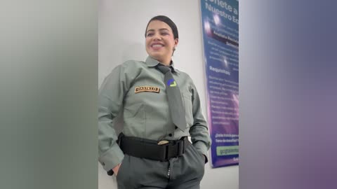 Video of a Latina woman with short dark hair, wearing a green \"Polic\u00eda\" uniform with badge and belt, standing in a clean, brightly lit room with informational posters in the background.