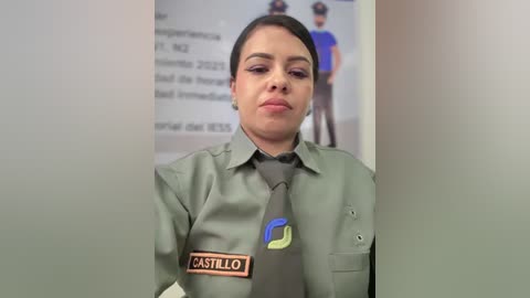 Video of a Latina woman in a green uniform with \"CASTILLO\" name tag, wearing dark makeup and a neutral expression. Background shows blurred office posters and another uniformed person.