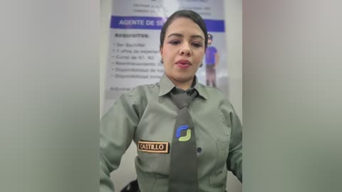 Video of a Latina woman with medium skin tone, dark hair, and full lips, wearing a green airport security uniform with a badge. She stands in front of a poster with Spanish text in a sterile airport environment.