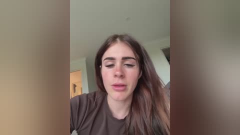Video of a young woman with long brown hair, wearing a dark shirt, sitting indoors with a neutral background, looking down with a neutral expression.