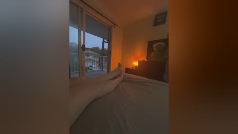 A video of a dimly lit bedroom with a person lying on a bed, covered by white sheets, beside a window with a balcony view.