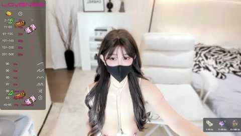 Video of a young Asian woman with long black hair, wearing a black face mask and revealing top, sitting on a white couch in a modern living room.