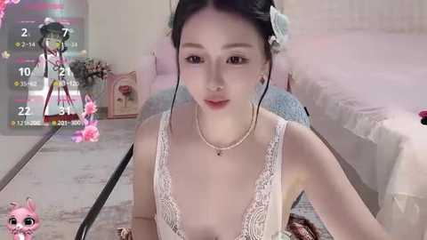 Video of an East Asian woman with fair skin, wearing a revealing white lace dress, sitting on a bed in a softly lit bedroom.