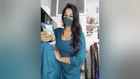 Video of a South Asian woman with long black hair, wearing a teal salwar kameez, gray face mask, and earrings, taking a selfie in a modern kitchen.