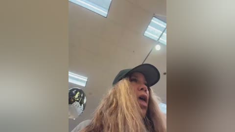 A low-angle video of a young woman with long blonde hair, wearing a black cap, exclaiming in surprise or joy, against a white ceiling with multiple rectangular skylights.