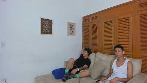 Video of two young men on a beige sofa, one wearing a black shirt and blue shorts, the other in a white tank top, in a cozy living room with wooden shutters and coffee-themed wall art.