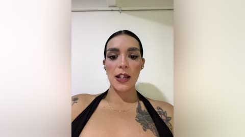 Video of a light-skinned woman with dark hair pulled back, wearing a black halter top, revealing tattoos on her shoulders, standing in a plain, white-walled room.