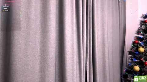 Video of a Christmas-themed room with a gray curtain partially covering a window, a lit Christmas tree adorned with red and gold ornaments on the right, and a corner of a white wall visible.