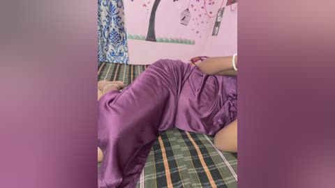 Video of a person lying on a green plaid bedspread, dressed in a purple skirt and shirt, with a pink wall featuring a tree painting and blue curtains in the background.