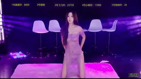 Video of an Asian woman with long, dark hair, wearing a sheer, nude-colored dress, standing in a dimly lit, modern room with white chairs and pink accents.