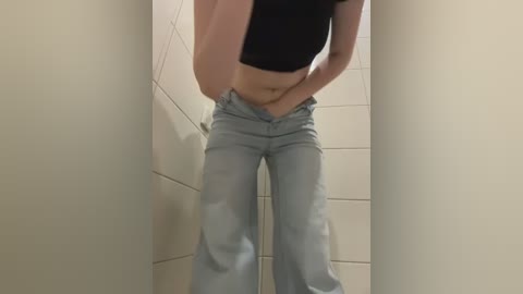 Media: Video of a light-skinned person with a slender build, wearing a black crop top and light blue jeans, standing in a narrow, tiled hallway.