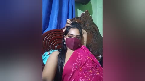A video shows a South Asian woman with medium-dark skin, wearing a red saree, mask, and long black hair, lying on a wooden bed with blue curtains, in a dimly lit room.