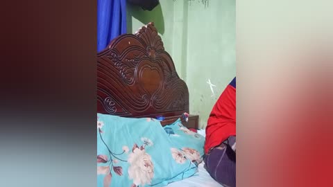 Video of a wooden bed with intricate carvings, blue floral sheets, and a red jacket partially visible, against a green wall with a cross symbol.