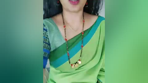 Video of a South Asian woman in a green and blue sari with intricate border, adorned with red and gold bead necklace, smiling with red lipstick.