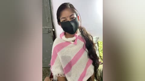 A video of a young woman with long black hair, wearing a pink and white striped crochet shawl, black face mask, and black top, in a dimly lit room with a grey door and green couch in the background.
