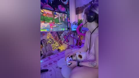 A video of a young woman with light skin, tattoos, and long dark hair, wearing a pink dress, playing a video game in a brightly lit, decorated room with neon lights and fairy lights.