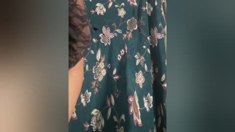 A video of a person wearing a dark green dress with a floral pattern featuring white, pink, and purple flowers. The person\u2019s arm, partially covered by a black lace sleeve, is visible on the left side. The background is blurred, focusing on the dress.