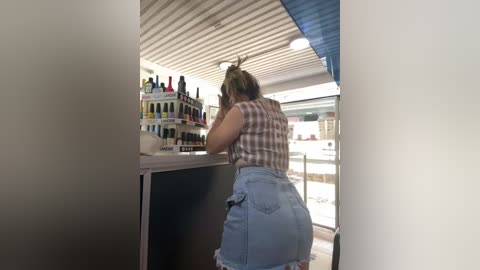 Video of a woman with light skin, medium build, and brown hair in a ponytail, wearing a plaid top and denim shorts, examining a display case filled with colorful lipsticks in a modern, brightly lit cosmetics store.