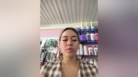 Video of a young woman with light skin and dark hair in a plaid shirt, standing in a pharmacy with shelves of cosmetics.
