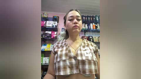 Video of a young Asian woman with long black hair, wearing a plaid crop top, standing in a brightly lit store filled with colorful cosmetics and skincare products.