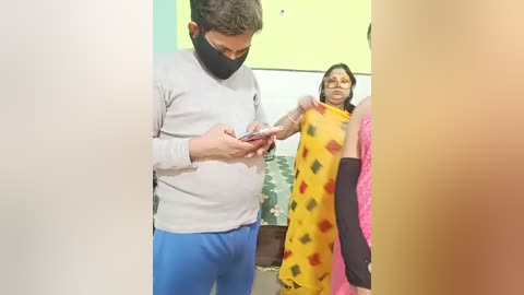 Video of a man in a grey sweater and blue pants, wearing a mask, examining a smartphone, standing next to a woman in a yellow sari with black and red patterns, and a woman in a pink sari. Background features green and yellow walls.
